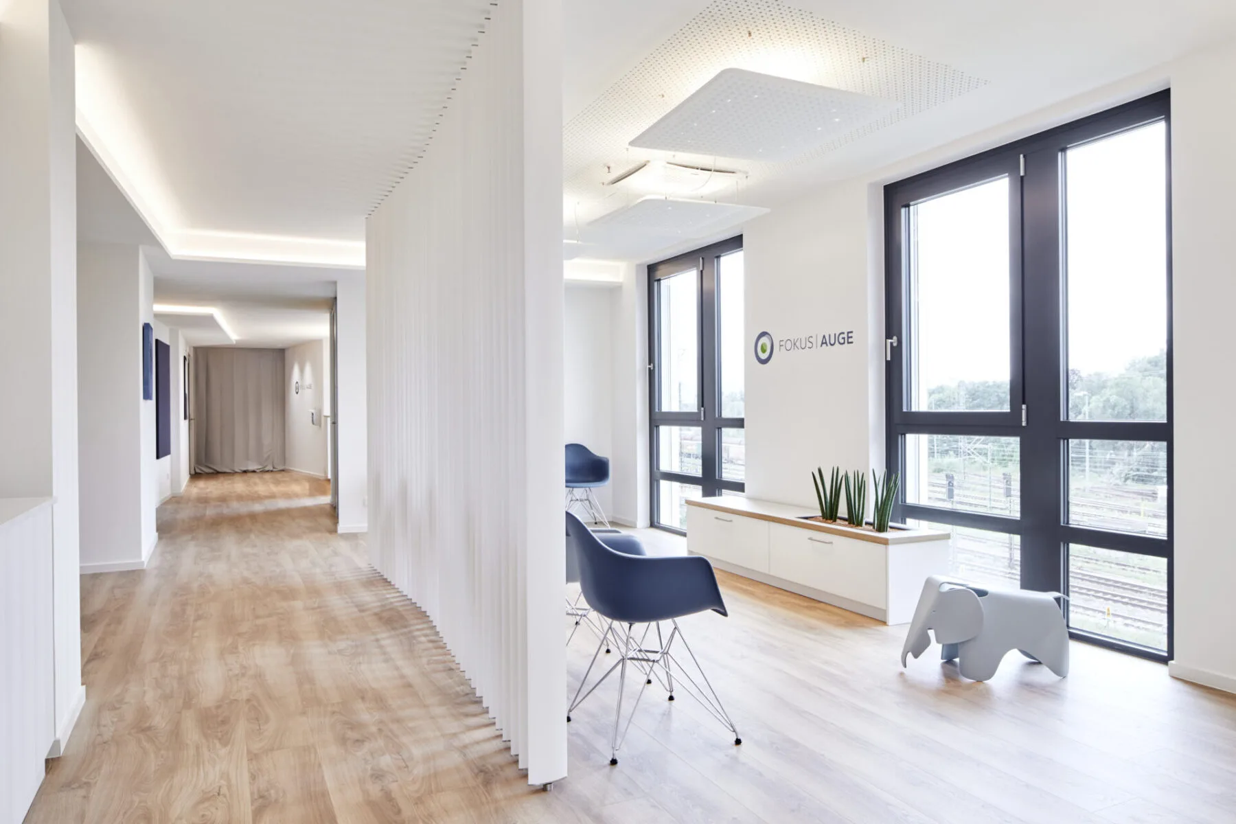 Blick durch den Praxiseingang mit Fensterfront, Wandlogo FOKUS AUGE, Sitzhocker und langem Flur