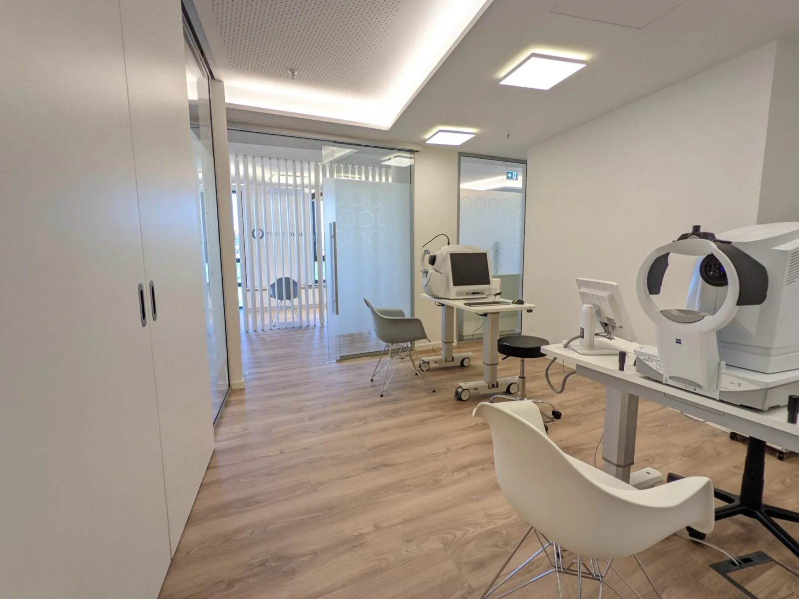 Untersuchungsraum mit großem augenärztlichem Messgerät, Tisch, Stuhl und Glasfront zum Flur